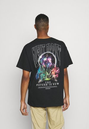 Persoon draagt een zwart T-shirt met een kleurrijk nevelontwerp, tekst "NIGHT ADDICT" en "THE FUTURE IS NOW" op de achterkant, tegen een effen achtergrond.