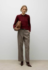 Woman standing wearing a burgundy long-sleeve polo, checkered trousers, dark pointed boots, large earrings, holding a brown leather bag under her arm.