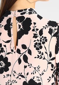Blusa rosa chiaro con motivo floreale nero, caratterizzata da un dettaglio con apertura a chiave sul retro e una chiusura con bottone al collo. Texture liscia.