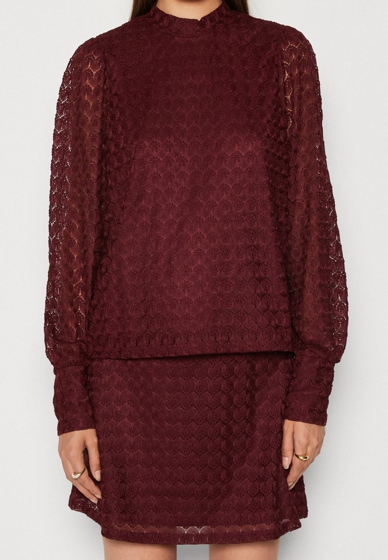 Robe en dentelle bordeaux avec manches longues bouffantes, col montant, poignets ajustés et mini-jupe assortie portée par une personne debout.