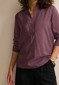 Blusa viola con scollo a V, maniche lunghe, sottile lucentezza e vestibilità rilassata, abbinata a pantaloni neri a righe e un braccialetto dorato.