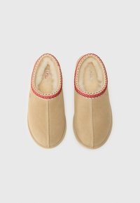 Beige pantoffels van zacht suede met een pluche interieur. Heeft een rode decoratieve stiksel langs de kraag en een ronde teenvorm.