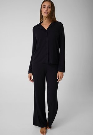 Femme debout pieds nus portant un haut noir à manches longues boutonné et un pantalon noir ample sur un fond gris uni.
