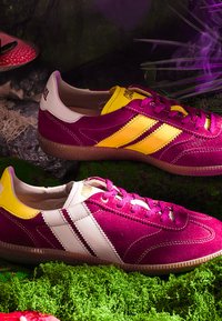 Deux baskets bordeaux avec des rayures jaunes et blanches reposant sur de la mousse verte avec un champignon rouge et une plante violette en arrière-plan.