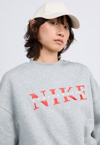 Sivi dukserica s crvenim izvezenim "NIKE" logotipom i bijelim natpisom "SPORTSWEAR". Mekani materijal, okrugli izrez, opušteni kroj, s suptilnom teksturom.