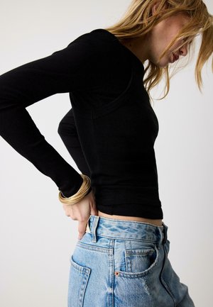 Ženska, ki si nadviguje svetlo modre kavbojke, oblečena v črn dolg rokav in več zlatih zapestnic, na belem ozadju.