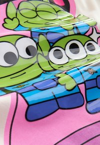 Graphic t-shirt featuring green cartoon aliens with large eyes, wearing blue outfits, printed on a pink background.