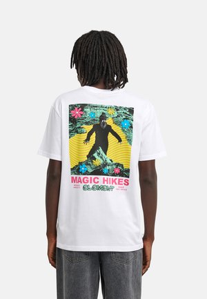 Personne portant un t-shirt blanc avec un graphisme coloré d'une silhouette barbu en randonnée, des fleurs et le texte "Magic Hikes Element" dans le dos.