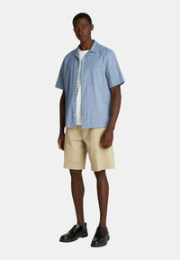 Camicia azzurra a maniche corte con bottoni, dotata di taschino sul petto, abbinata a pantaloni corti beige e scarpe nere con lacci. Tessuto strutturato.