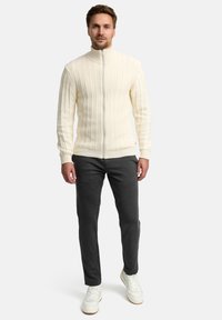 Cremefarbener Reißverschluss-Pullover mit geripptem Muster, kombiniert mit dunkelgrauer Hose und weißen Sneakern. Der Pullover hat einen hohen Kragen und ein figurbetontes Design.