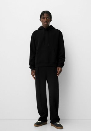 Schwarzer Kapuzenpullover mit Fronttasche, kombiniert mit weit geschnittenen schwarzen Hosen. Minimalistisches Design mit weichem Stoff und ohne sichtbare Muster.