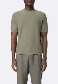T-shirt verde oliva a maniche corte con collo rotondo a costine, realizzato in un tessuto strutturato, abbinato a pantaloni in lino grigio chiaro.