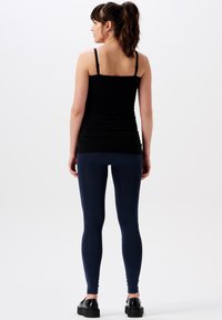 Kvinna med brunt hår i hästsvans iförd en svart linne, marinblå leggings och svarta platåskor, stående med ryggen vänd mot kameran på en vit bakgrund.