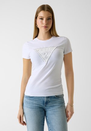 Donna che indossa una T-shirt bianca aderente con logo Guess in rilievo e jeans azzurri, in piedi contro uno sfondo chiaro e uniforme.