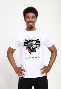 T-shirt bianco in cotone con una grafica di testa di mucca, accentuata da dettagli rossi e blu, e la scritta "Tâche de vache" sottostante.