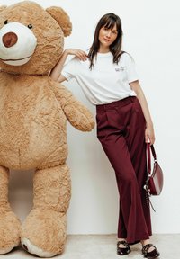Un ours en peluche marron avec une texture douce à côté d'une personne portant un t-shirt blanc, un pantalon bordeaux et des chaussures noires, tenant un sac à main bordeaux.