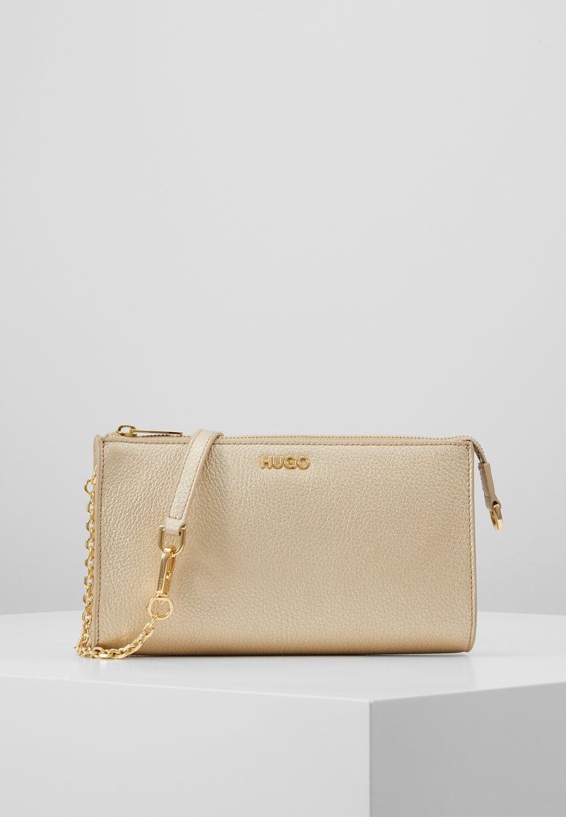 Złota torebka skórzana o teksturowanej powierzchni z łańcuszkowym paskiem, z zamknięciem na zamek i wytłoczonym logo "HUGO" z przodu. Kompaktowy prostokątny kształt.