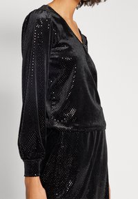 Svart sammetsöverdel med V-ringning, prydd med ett prickigt mönster, puffärmar och lös passform. Slät textur med en subtil lyster.