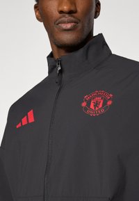 Μαύρο μπουφάν με ψηλό γιακά, που φέρει το κόκκινο λογότυπο της Manchester United και τρεις κόκκινες ρίγες στην αριστερή πλευρά. Κατασκευασμένο από ελαφρύ υλικό.