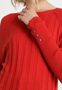 Pull côtelé rouge avec un col rond et des boutons décoratifs sur le poignet. Le tissu semble texturé et moulant.