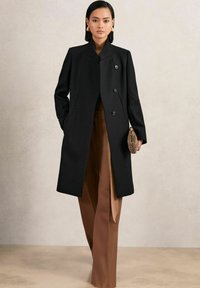 Schwarzer maßgeschneiderter Mantel mit hochstehendem Kragen, der drei Knöpfe auf der Vorderseite hat. Trägt über braunen Hosen und zeigt eine elegante Silhouette.
