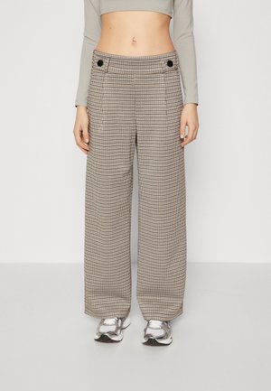 Kobieta w beżowo-czarne, wysokowalne szerokie spodnie w kratę, szary długi crop top i srebrne sneakersy, stojąca na jednolitym tle.