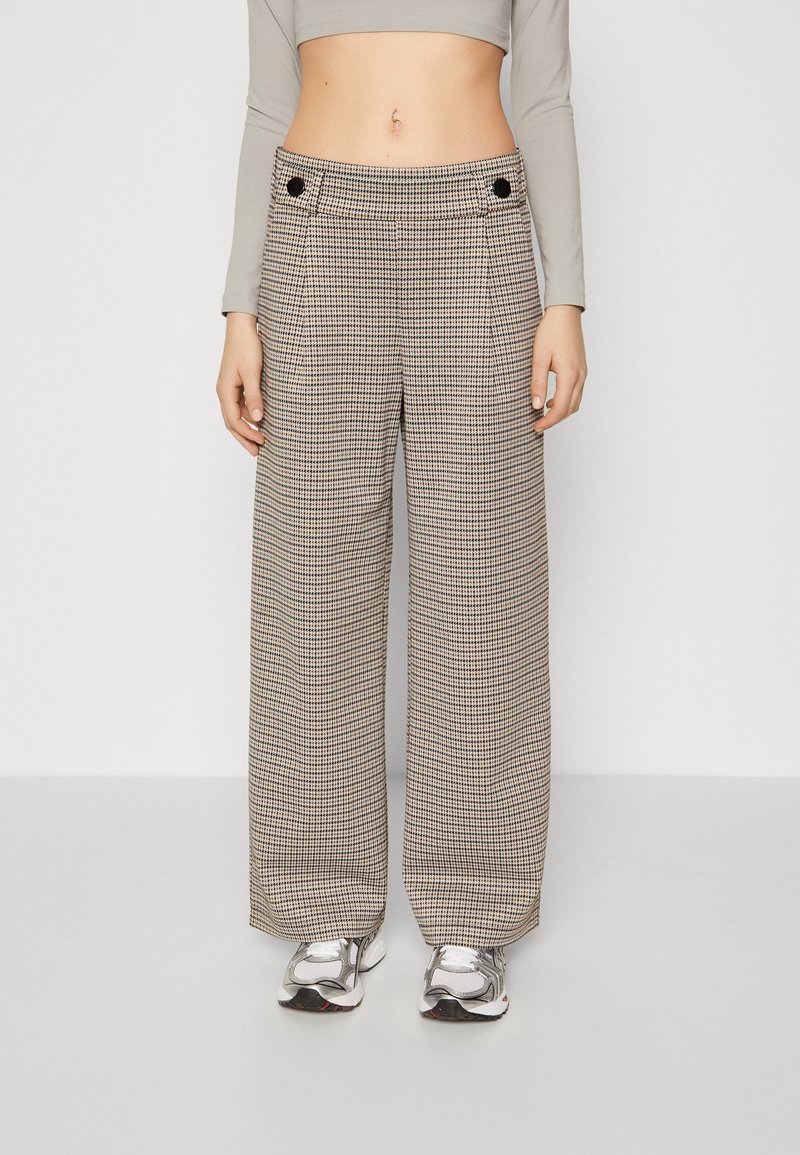 Femme portant un pantalon beige et noir à carreaux, taille haute, jambes larges, un crop top gris à manches longues et des baskets argentées, debout devant un fond uni.