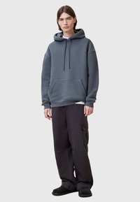 Felpa grigia con cappuccio, dotata di tasca frontale e lacci, abbinata a pantaloni cargo neri e scarpe slip-on nere. Design semplice, vestibilità casual.