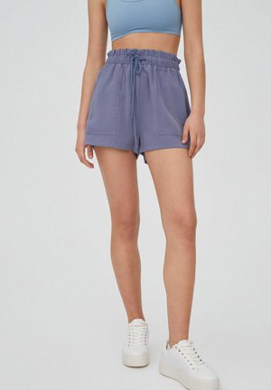 Femme portant un short taille haute bleu à cordon avec des poches avant, un crop top bleu clair et des baskets blanches à plateforme sur un fond uni.