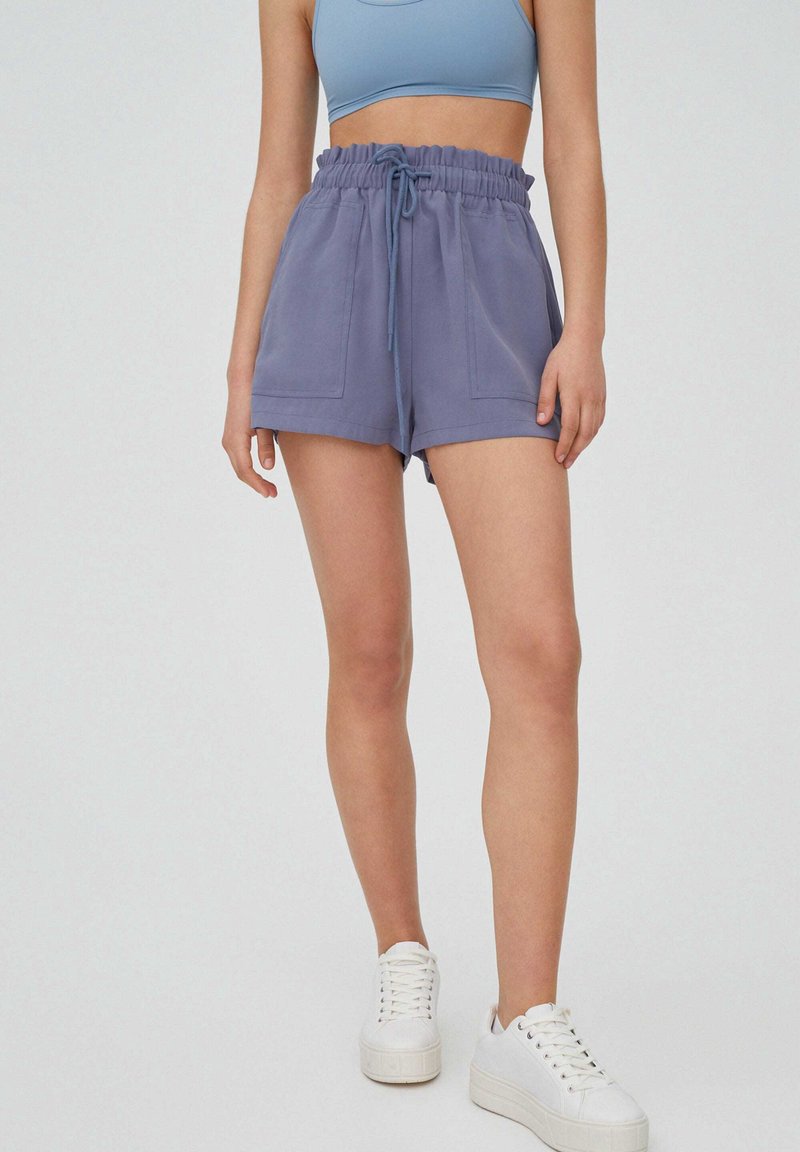 Femme portant un short taille haute bleu à cordon avec des poches avant, un crop top bleu clair et des baskets blanches à plateforme sur un fond uni.