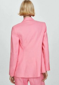 Persona rubia de espaldas, vistiendo una chaqueta de traje rosa brillante a medida y pantalones a juego, contra un fondo claro y liso.