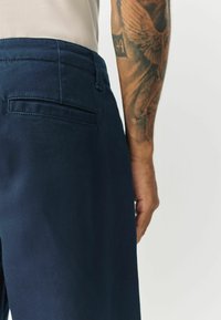 Navy blue trousers with a smooth texture, featuring a back pocket and subtle stitching details. Visible tattoo on the forearm.