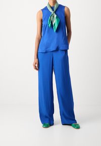 Haut bleu sans manches et pantalon à jambes larges en tissu doux. Accessoirisé d'une écharpe verte et blanche à motifs et de chaussures plates vertes.