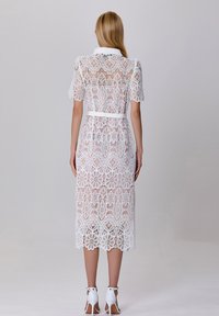 Robe en dentelle blanche à manches courtes, motif floral et taille ceinturée. L'ourlet a un bord festonné. Portée avec des talons blancs.