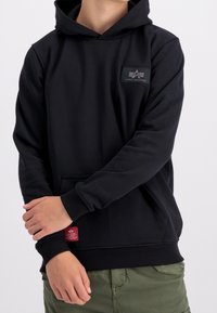 Felpa nera con cappuccio realizzata in tessuto morbido, con una tasca frontale, polsini a costine e un'etichetta con il marchio sul petto, abbinata a pantaloni color oliva.