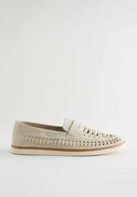 Scarpa slip-on beige chiaro realizzata in materiale morbido, con un design superiore intrecciato, suola piatta e una banda di accento marrone chiaro attorno alla base.