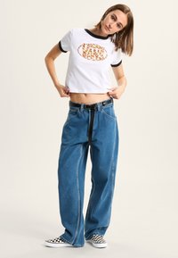White cropped t-shirt with black ringer neckline and a leopard-print Vans logo; loose blue jeans with black belt; checkered slip-on shoes.