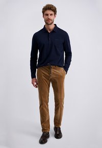 Navy long-sleeve polo with a collar and front pocket, paired with brown corduroy pants. Model wears dark shoes, standing against a light background.