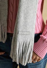 Sivá pletená šál s strapcami na koncoch, ozdobený záplátkou s logom. Nosí sa cez ružový rebrovaný sveter. Texturovaná tkanina s mäkkým vzhľadom.