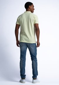 Ljusgul polo skjorta med korta ärmar, ihopparad med åtsittande blå jeans och vita sneakers, sedd bakifrån mot en enfärgad bakgrund.