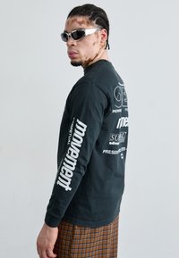 Black long-sleeve shirt with white graphic text on sleeves and back. Patterned brown plaid pants. Individual wearing sunglasses, facing sideways.