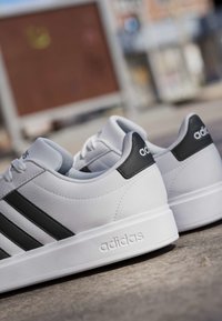 Weiße Adidas-Sneaker mit schwarzen Streifen und Fersenlasche, die im Freien auf einer Betonoberfläche platziert sind.