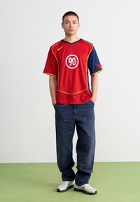 Jersey deportivo rojo y azul marino con un logo redondo que presenta el número "90". Unos vaqueros azules holgados y zapatillas blancas completan el conjunto.