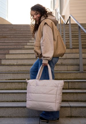 Ung kvinde med krøllet hår går op ad udendørs trapper med en stor beige quiltet skuldertaske og iført lag-på-lag afslappet tøj.