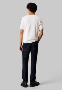 Camiseta blanca de manga corta, jeans de denim azul y zapatos negros. Los jeans tienen un diseño clásico de cinco bolsillos y costuras visibles en la parte trasera.