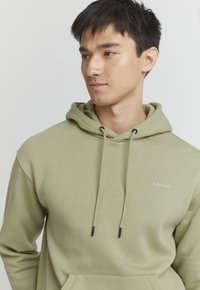 Olivovo zelená mikina so soft materiálom, obsahuje predné vrecko, šnúrky s čiernymi koncami a logo "blend" v bielej farbe na hrudi.
