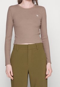 Maglione lungo a maniche sia in marrone con scollo a girocollo, caratterizzato da un piccolo emblema ricamato. Abbinato a pantaloni verdi oliva a vita alta.