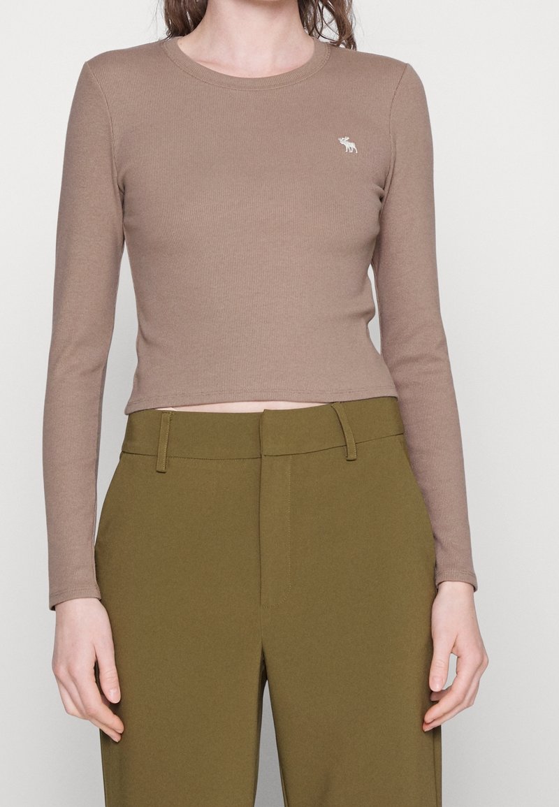 Maglione lungo a maniche sia in marrone con scollo a girocollo, caratterizzato da un piccolo emblema ricamato. Abbinato a pantaloni verdi oliva a vita alta.