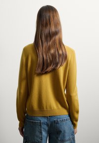 Pullover senape realizzato in tessuto morbido, con una vestibilità comoda e orlo a costine, abbinato a jeans blu chiaro a vita alta.