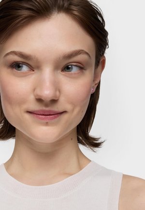 Młoda kobieta z krótkimi brązowymi włosami, ubrana w biały top bez rękawów, patrząca na bok z lekkim uśmiechem na jednolitym tle.